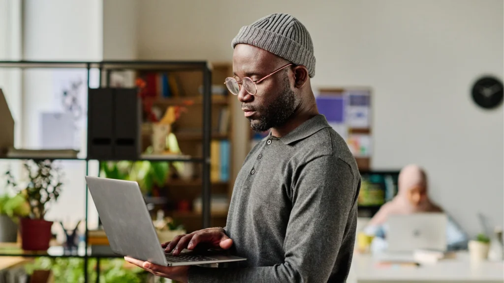 A young black man, holding his laptop as he works in an office.